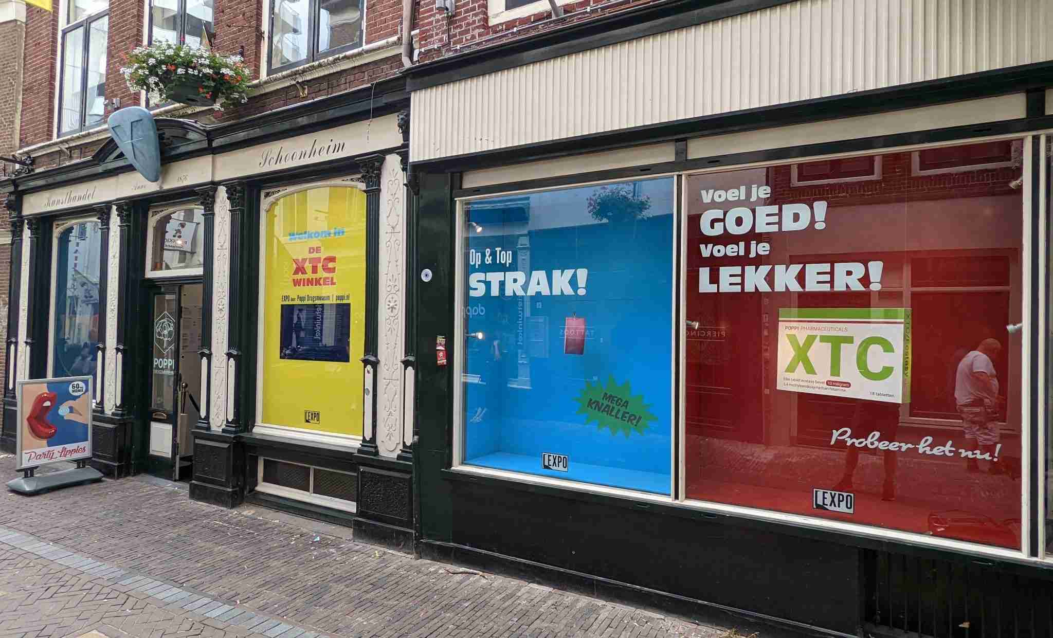 Una tienda en una calle adoquinada con una fachada tradicional y un toldo azul. En las ventanas se observan carteles coloridos que hacen referencia al XTC. Un cartel amarillo dice 