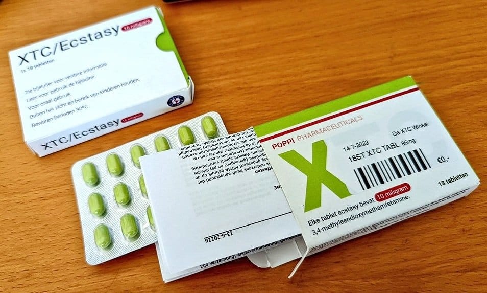 Empaque de tabletas de XTC/Ecstasy producido por Poppi Pharmaceuticals. La caja muestra información detallada del producto, incluyendo la dosis de 10 miligramos de 3,4-metilendioximetanfetamina por tableta y fecha de caducidad. Se visualizan pastillas verdes en un blíster y detalles de etiquetas e instrucciones en varios lados de la caja.