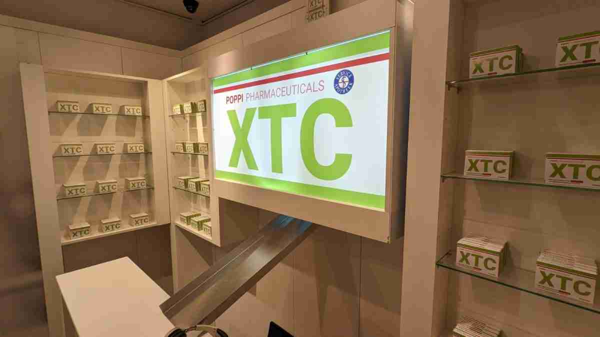 Una imagen muestra un espacio interior con estanterías blancas que exhiben cajas etiquetadas como "XTC". Hay un mostrador blanco en la parte inferior. Sobre las estanterías hay un letrero luminoso que dice "Poppi Pharmaceuticals XTC" con un logo en la parte superior izquierda.