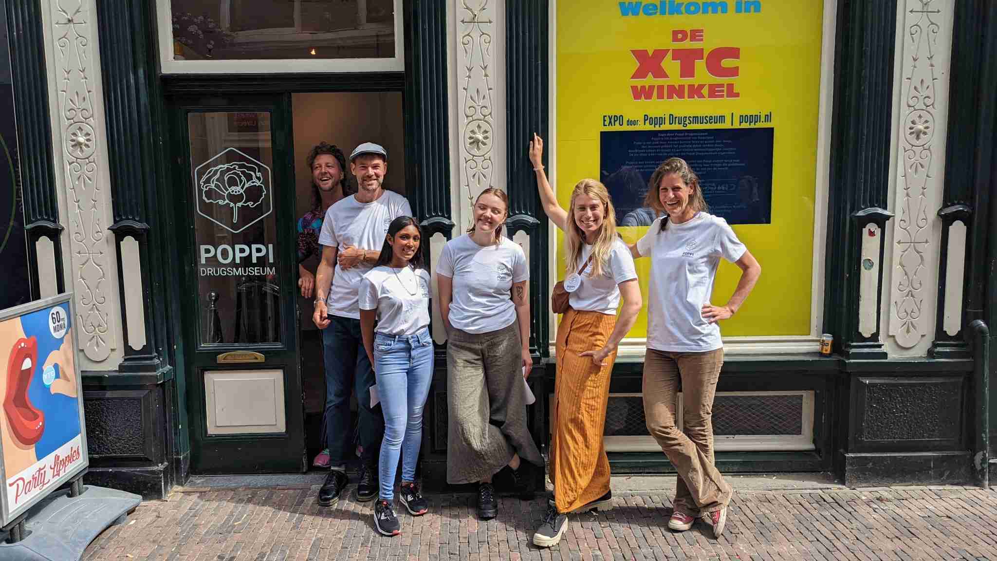 Un grupo de personas vestidas con camisetas blancas con el logotipo de Poppi posan frente a una entrada decorada con detalles arquitectónicos tradicionales. Se destaca un cartel amarillo que dice 'DE XTC WINKEL' junto a la puerta del 'Poppi Drugsmuseum'. Las personas exhiben sonrisas amistosas y muestran orgullo y camaradería.