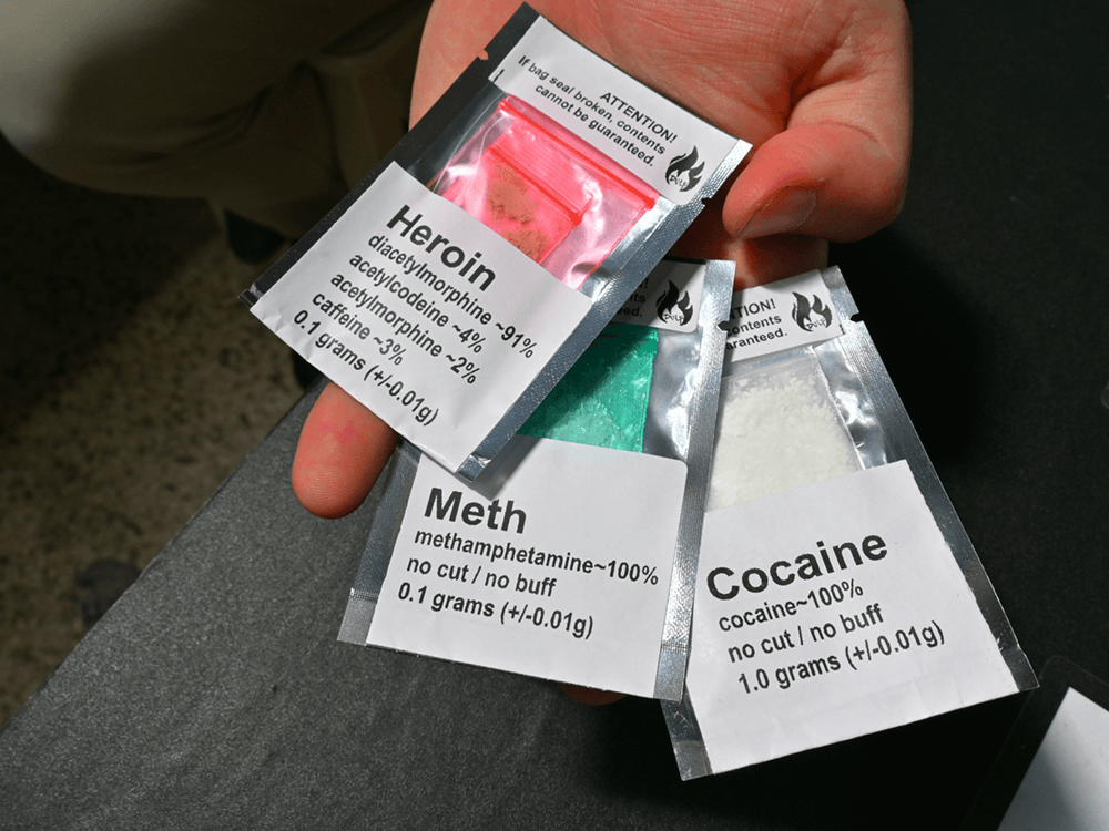 Una mano sostiene tres bolsas pequeñas y transparentes, etiquetadas individualmente con los nombres de diferentes sustancias: "Heroin", "Meth", y "Cocaine". Cada bolsa contiene polvos de diferentes colores y texturas, correspondientes a la sustancia señalada. Las etiquetas también proporcionan información sobre la pureza y la cantidad de cada sustancia en gramos, con la heroína siendo la única que tiene sustancias adicionales listadas. Las advertencias de "flammability" (inflamabilidad) aparecen en la parte superior de cada bolsa, señalando la naturaleza potencialmente peligrosa de los contenidos.
